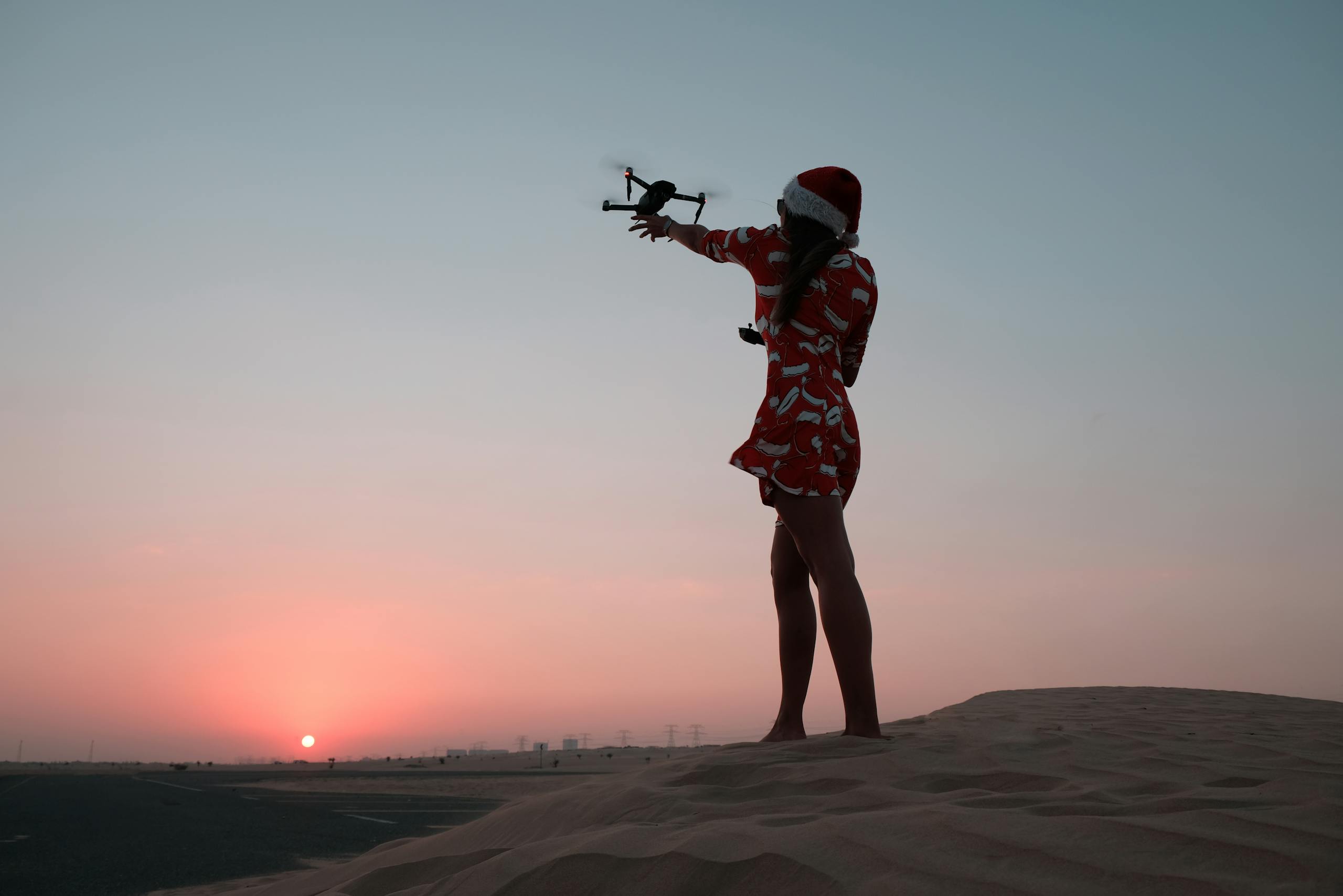 A woman in a Santa hat controls a drone on desert dunes at sunset.